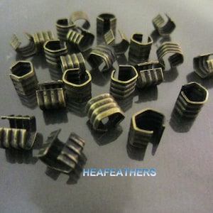 May include: A close-up of a group of antique gold metal crimp beads. The beads are small and have a hexagonal shape with a rounded edge. The beads are arranged in a random pattern on a grey surface.