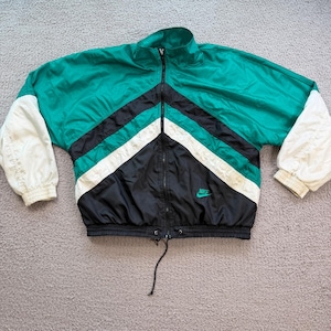 May include: A zip-up windbreaker jacket featuring a color-blocked design in emerald green, black, and white. The jacket has long white sleeves with elastic cuffs, a high collar, and a black drawstring hem. A small green Nike logo is on the chest.