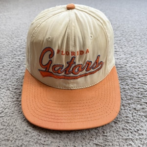May include: A tan and orange baseball cap with the word "FLORIDA Gators" embroidered in orange script. The cap has a curved brim and a button on top.