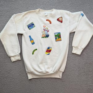 May include: Cream-colored sweatshirt with multiple embroidered patches. Patches include a bottle, a can, and a cartoon character with a dog. Other patches have the words "CONSERVE" and "PROTECT".