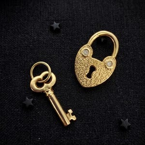 May include: A gold-toned heart-shaped padlock with a textured surface and two small embedded clear stones, accompanied by a matching gold-toned key with a circular ring top, both displayed against a dark black fabric background.