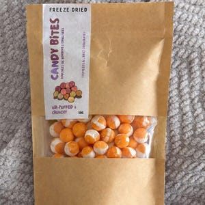May include: A brown paper bag with a clear window displaying orange and white freeze-dried candy. The bag has a label that reads "CANDY BITES" and "AIR-PUFFED & CRUNCHY". The bag contains 50g of candy.