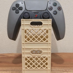 May include: A gray and black PlayStation 5 controller is displayed above two stacked, cream-colored plastic crates. The controller features a PlayStation logo and colored button symbols. The crates have a lattice design.