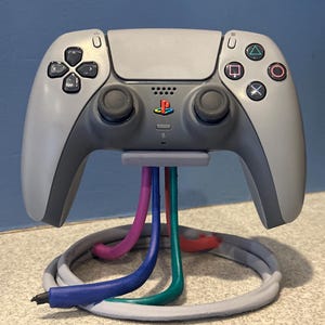 May include: A gray PlayStation 5 controller on a matching stand with colorful, stylized cables. The stand's base is a gray loop. The controller has black buttons and the PlayStation logo.