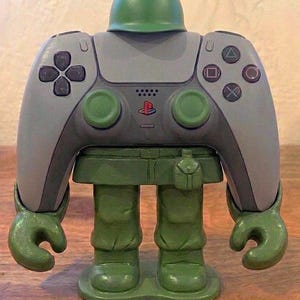 Op de afbeelding: Een grijze videogamecontroller geïntegreerd in een groen plastic soldaatfiguur. De controller is voorzien van een richtingspad, vier actieknoppen, twee analoge sticks en een PlayStation-logo. Het figuur heeft een helm, armen en benen.
