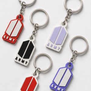 May include: Five keychains featuring a shield-shaped pendant with the word ARMY embossed at the bottom. The pendants come in various colour combinations: red with white, white with black, white with light purple, white with red, and purple with white.