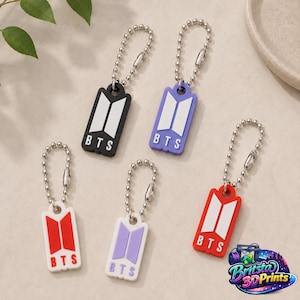 Puede incluir: Cinco llaveros rectangulares con cadenas de bolas plateadas, cada uno con el logotipo y el texto BTS en blanco. Los llaveros vienen en negro, morado, blanco con logo rojo, lila y rojo con logo blanco, dispuestos sobre una superficie neutra.