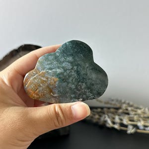 Puede incluir: Una escultura de piedra con forma de nube, con tonos verdes, marrones y blancos. La piedra pulida se sostiene en una mano. El objeto decorativo es una pieza única.