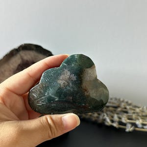 Puede incluir: Una piedra en forma de nube con una mezcla de colores verdes, marrones y blancos. La superficie pulida refleja la luz, resaltando los patrones naturales dentro de la piedra. La piedra está sostenida en una mano.
