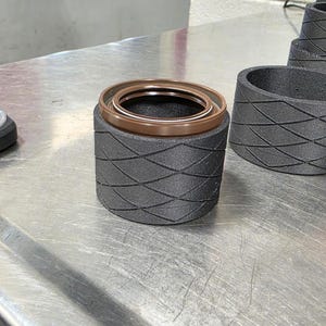 Op de afbeelding: Close-up van een cilindrische component met een getextureerde, donkergrijze buitenkant en een bruine binnenring. De component staat op een metalen oppervlak, met meer soortgelijke componenten op de achtergrond.