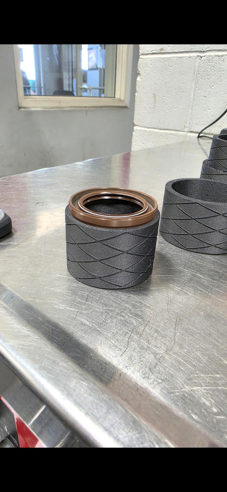Pode incluir: Um selo mec&acirc;nico cil&iacute;ndrico com um exterior cinza-escuro texturizado com um padr&atilde;o de diamante repetitivo e uma inser&ccedil;&atilde;o circular marrom lisa no topo. O componente tem uma apar&ecirc;ncia industrial r&iacute;gida com um anel met&aacute;lico interno vis&iacute;vel.