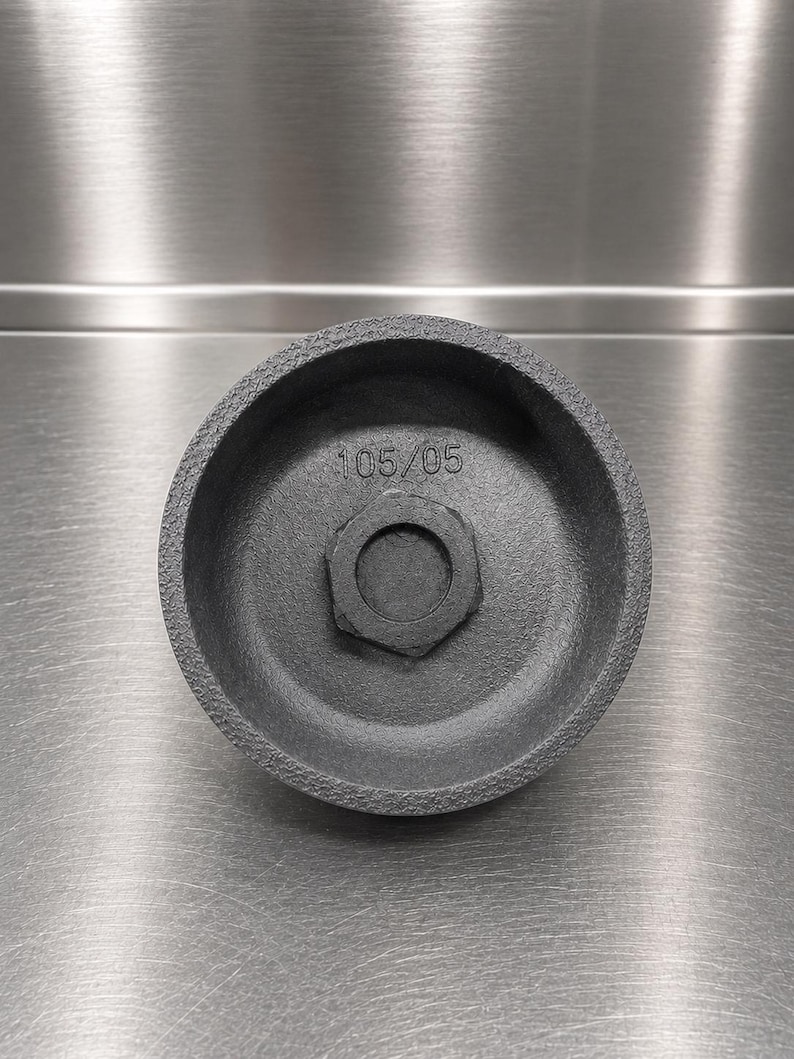 Pode incluir: Uma chave de filtro de &oacute;leo circular de metal cinza-escuro com acabamento exterior texturizado. O interior apresenta uma porca hexagonal central e o texto estampado 105/05. A ferramenta tem uma forma profunda de copo para manuten&ccedil;&atilde;o.
