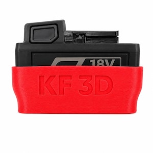 Pode incluir: Uma bateria de ferramenta elétrica de 18V preta dentro de uma capa protetora vermelha brilhante. A capa vermelha apresenta o texto em relevo KF 3D na parte frontal. A bateria tem uma carcaça de plástico preto com um botão de liberação no topo.
