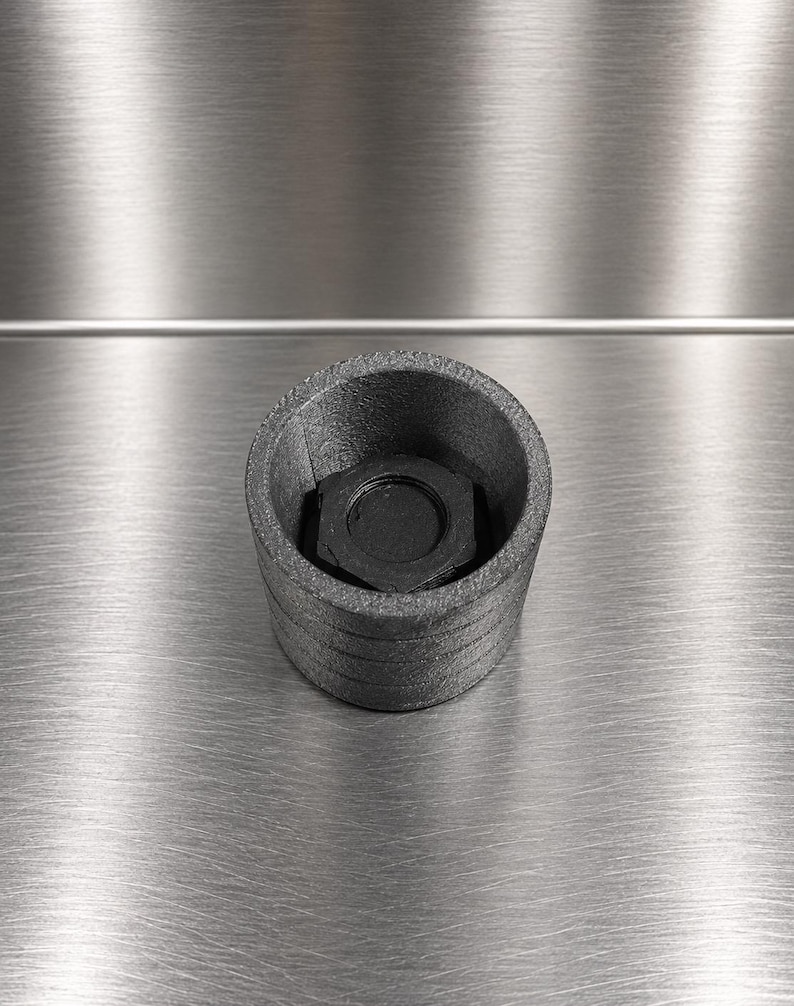 Pode incluir: Uma caixa cil&iacute;ndrica de pl&aacute;stico cinzento-escuro texturizado com um centro oco contendo uma porca hexagonal preta. A superf&iacute;cie exterior apresenta sulcos horizontais subtis e o componente &eacute; mostrado a partir de uma perspetiva superior.