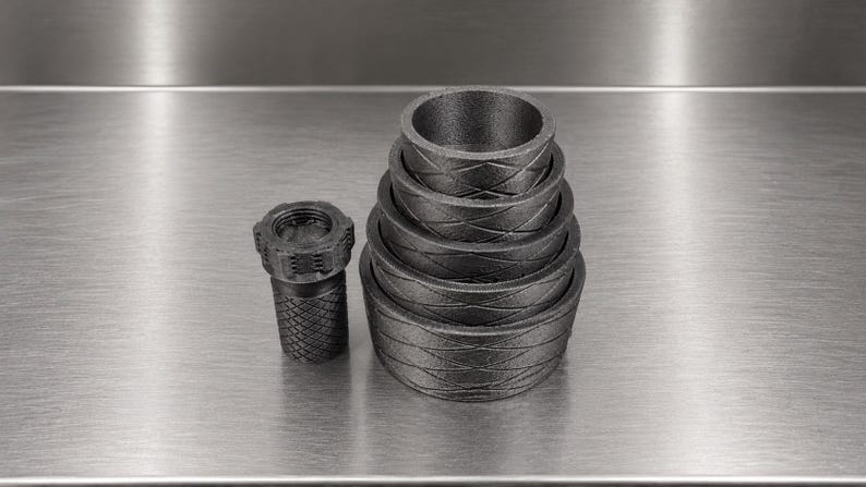 Pode incluir: Um conjunto de cinco an&eacute;is circulares cinza-escuro encaixados com um padr&atilde;o de diamante texturizado no exterior, acompanhado por um componente cil&iacute;ndrico &uacute;nico com uma textura de diamante correspondente e uma parte superior roscada.