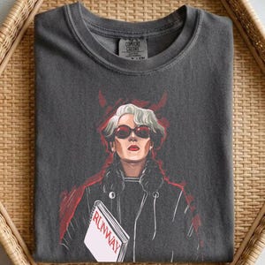 Puede incluir: Camiseta gris oscuro con una ilustración estilizada de una mujer con gafas de sol rojas y una chaqueta negra, sosteniendo un libro con la palabra "RUNWAY". Marca Comfort Colors.