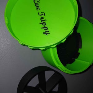May include: Bright green container with a lid, featuring the text "Live Trippy" in black script. The container is open, revealing a dark interior. A black, circular object with spokes is also visible.