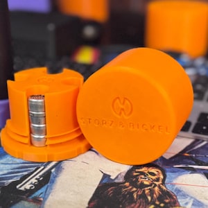 May include: Bright orange vaporizer accessories. One piece is cylindrical with the text "STORZ & BICKEL" and a logo. The other piece is a disassembled orange component with silver metal parts. The items are on a patterned surface.