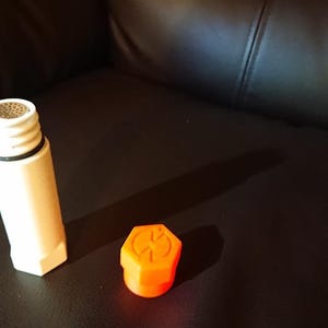 May include: An off-white, hexagonal object with a mesh top and a separate bright orange, hexagonal cap with a symbol. The objects are on a dark brown surface. The cap has a symbol on it.