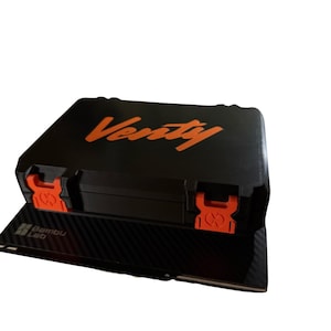 Może przedstawiać: Czarna walizka z pomarańczowymi detalami i napisem "Verity" w kolorze pomarańczowym. Walizka ma dwa pomarańczowe zatrzaski i stoi na czarnej powierzchni z logo "Bambu Lab".