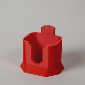 Puede incluir: Un soporte rojo brillante, impreso en 3D, con un diseño geométrico. El soporte presenta una base rectangular con forma hexagonal, un compartimento central y una pequeña extensión rectangular en la parte superior. El objeto está hecho de un material sólido y opaco.