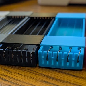May include: A close-up of several small, rectangular, plastic containers in various colours, including grey, black, blue, and orange. Each container has a series of metal hooks attached to the front. The containers are arranged side by side on a wooden surface.