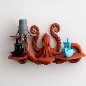 May include: A 3D-printed octopus-shaped shelf in a reddish-brown color. The shelf has three platforms, each holding a different object: a gray figure with "10mm" printed on it, a black boat, and a blue boat. The octopus's tentacles curl upwards.