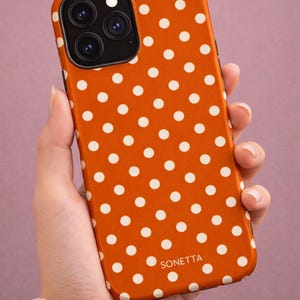 May include: Orange phone case with a white polka dot pattern. The case has a glossy finish and features the brand name "SONETTA" at the bottom. The phone's camera lenses are visible.