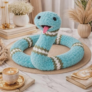 Peut inclure: Un serpent en peluche fabriqué en fil bleu clair doux avec une finition texturée. Le serpent présente des yeux en plastique noir, une langue en feutrine rose et des bandes horizontales de motifs en fil blanc et vert olive le long du corps.