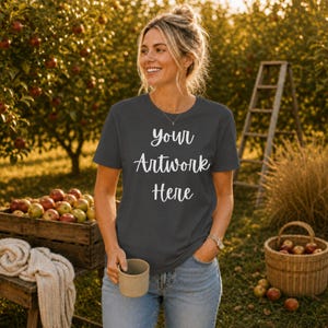 May include: A dark gray short-sleeved t-shirt featuring the white text "your Artwork Here" printed on the front. The shirt has a crew neckline and is worn with blue denim jeans. A plain, light-colored ceramic mug is held in one hand.