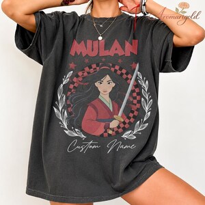 May include: Dark grey t-shirt with the word "MULAN" in red above a graphic of Mulan holding a sword. The graphic is set within a checkered pattern and a white floral wreath. The words "Custom Name" are written below the graphic in a script font.