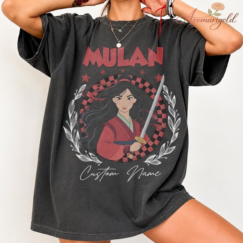 May include: Dark grey t-shirt with the word "MULAN" in red above a graphic of Mulan holding a sword. The graphic is set within a checkered pattern and a white floral wreath. The words "Custom Name" are written below the graphic in a script font.