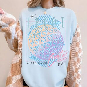 May include: Light blue t-shirt featuring a colorful graphic of a geometric sphere with the text "EPCOT Center" and "Walt Disney World 1982". The design includes wavy lines and stars, creating a retro aesthetic.