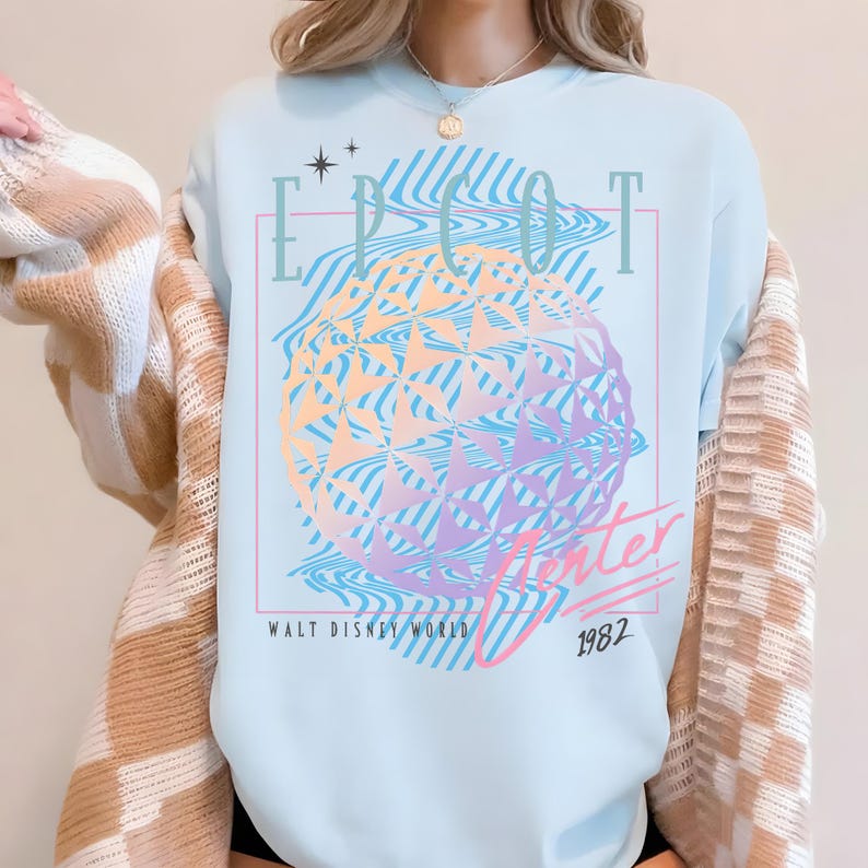 May include: Light blue t-shirt featuring a colorful graphic of a geometric sphere with the text "EPCOT Center" and "Walt Disney World 1982". The design includes wavy lines and stars, creating a retro aesthetic.