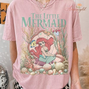 May include: Pink t-shirt featuring a graphic of Ariel from The Little Mermaid, seated in a seashell. The design includes the text "The Little Mermaid" in teal, with coral, fish, and other sea elements.
