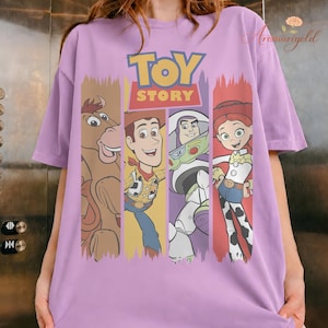 Puede incluir: Una camiseta lila con el logo de Toy Story y cuatro personajes: Bullseye, Woody, Buzz Lightyear y Jessie. Los personajes se muestran en paneles verticales con un efecto de pintura desgastada. La camiseta es de material suave.