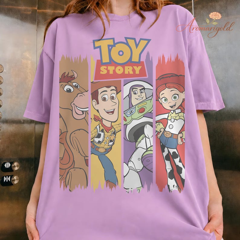 Puede incluir: Una camiseta lila con el logo de Toy Story y cuatro personajes: Bullseye, Woody, Buzz Lightyear y Jessie. Los personajes se muestran en paneles verticales con un efecto de pintura desgastada. La camiseta es de material suave.