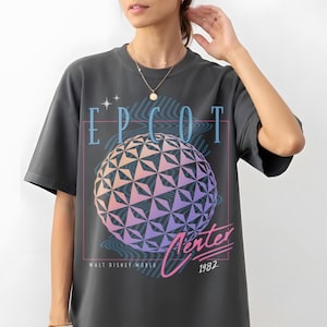May include: Dark gray t-shirt featuring a graphic of the EPCOT Center sphere with text that reads "EPCOT Center Walt Disney World 1982". The graphic is in shades of blue, pink, and purple.