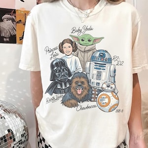 May include: Cream-colored t-shirt featuring a Star Wars graphic. The design includes illustrations of Darth Vader, Princess Leia, Baby Yoda, Chewbacca, R2-D2, and BB-8, with their names above or below them. The shirt is a casual wear item.