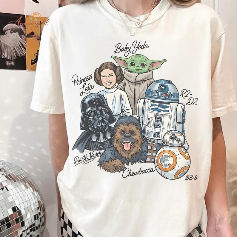 May include: Cream-colored t-shirt featuring a Star Wars graphic. The design includes illustrations of Darth Vader, Princess Leia, Baby Yoda, Chewbacca, R2-D2, and BB-8, with their names above or below them. The shirt is a casual wear item.