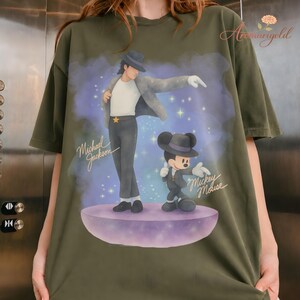 May include: Olive green t-shirt featuring a graphic of Michael Jackson and Mickey Mouse in a stylized design. The text "Michael Jackson" and "Mickey Mouse" are also visible. The shirt has short sleeves and a relaxed fit.