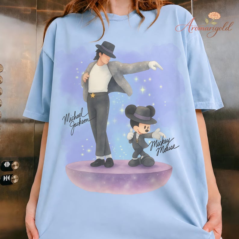 May include: Light blue t-shirt featuring a graphic of Michael Jackson and Mickey Mouse in matching outfits. The design includes the text "Michael Jackson" and "Mickey Mouse" in cursive script. The illustration is set against a purple and blue background.