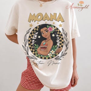 May include: Cream-coloured t-shirt with the word "MOANA" in gold block letters above a circular graphic. The graphic features a cartoon illustration of a woman with flowers in her hair, surrounded by a black and white checkered pattern and floral elements.