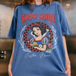 May include: Blue t-shirt with a Snow White graphic and the text "SNOW WHITE" in red. The design includes a checkered circle, floral elements, and the words "Custom Name".