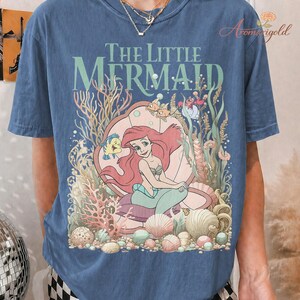 May include: Blue t-shirt featuring a graphic of Ariel from The Little Mermaid. The design includes Ariel, Flounder, and other sea creatures, with the words "The Little Mermaid" above the image. The shirt has a vintage, faded look.