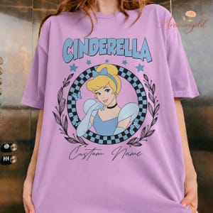 May include: A lilac t-shirt with a Cinderella graphic. The design features the word "CINDERELLA" in light blue, a portrait of Cinderella, and the words "Custom Name". The graphic is framed by a black and white checkered circle and a floral wreath.