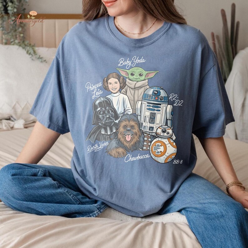 May include: Blue t-shirt featuring a graphic of Star Wars characters including Baby Yoda, Princess Leia, Darth Vader, Chewbacca, R2-D2, and BB-8. The text "Baby Yoda", "Princess Leia", "Darth Vader", "Chewbacca", "R2-D2", and "BB-8" are also included.