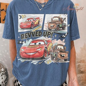 May include: Blue t-shirt featuring a comic strip design with Lightning McQueen and Mater from the Cars movie. The phrase "REVVED UP!" is printed in white. The shirt has a crew neck and short sleeves.