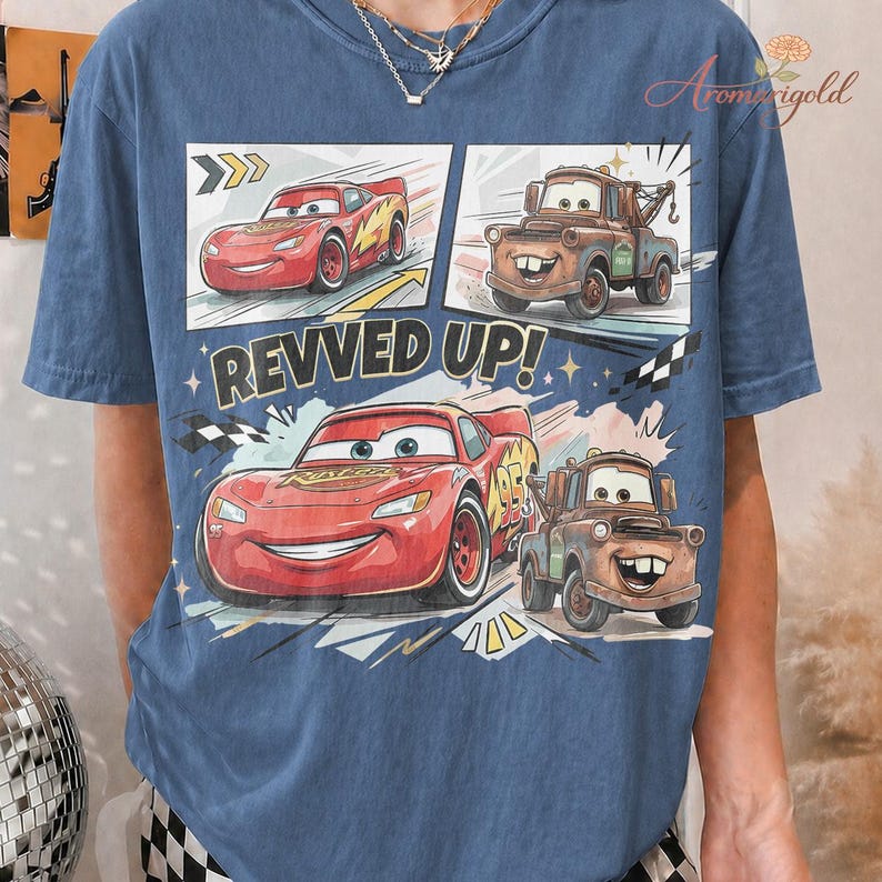 May include: Blue t-shirt featuring a comic strip design with Lightning McQueen and Mater from the Cars movie. The phrase "REVVED UP!" is printed in white. The shirt has a crew neck and short sleeves.