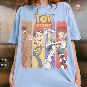 Puede incluir: Camiseta azul claro con las palabras "TOY STORY" en amarillo y rojo, y personajes de dibujos animados. El dise&ntilde;o incluye un caballo marr&oacute;n, Woody, Buzz Lightyear y Jessie, cada uno en un bloque de color vertical.